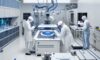 Engineering students in white cleanroom suits inspect a silicon wafer under a microscope in a high-tech semiconductor fabrication lab with robotic arms in the background.