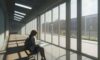 A student sits on a bench in a sunlit, modern high school hallway, focused intensely on a glowing laptop screen displaying a complex AI interface.