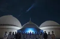 A group of diverse Indian students holding certificates and wearing badges, standing proudly in front of a large observatory dome under a starry night sky.
