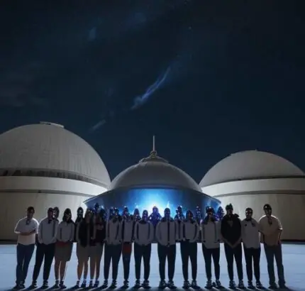 A group of diverse Indian students holding certificates and wearing badges, standing proudly in front of a large observatory dome under a starry night sky.