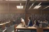 A student sits at a large wooden table in a modern university library, studying with textbooks, a laptop, and a cup of coffee amidst rows of books.
